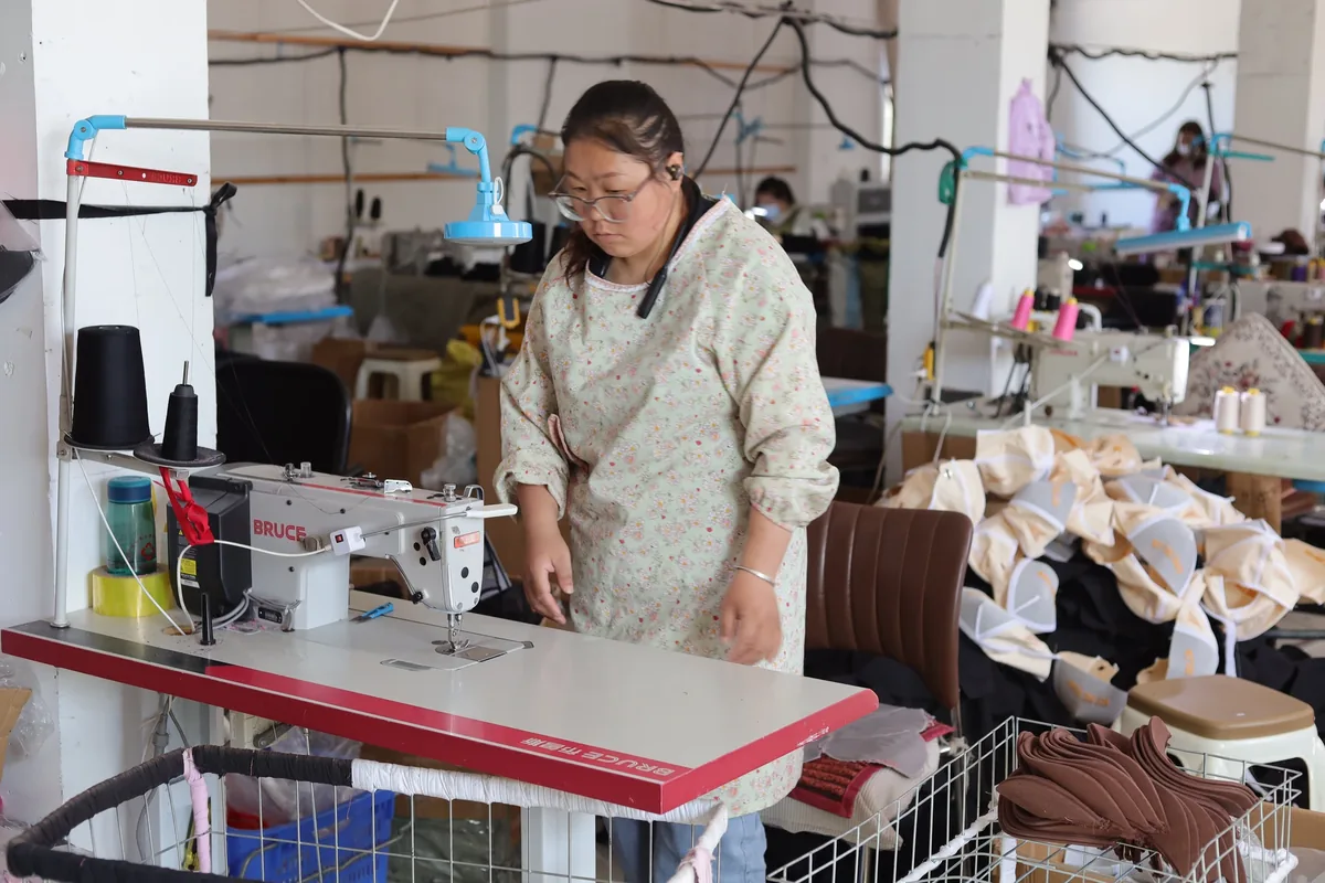 An operator beside a sewing machine with white cap components and stacked brown pieces nearby