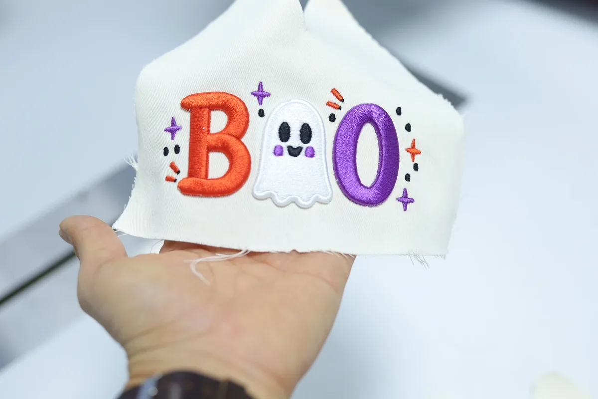 A hand holding a white fabric panel with orange, purple and white embroidery